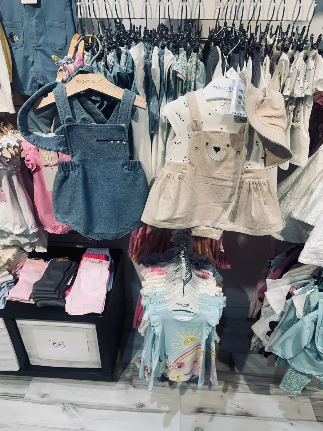 Percheros con ropa de bebé y de niña: un peto vaquero y un pichi color crema con una cara de oso.